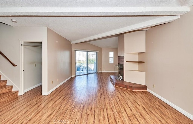 unfurnished living room featuring baseboards, a fireplace with raised hearth, lofted ceiling with beams, stairs, and light wood-type flooring