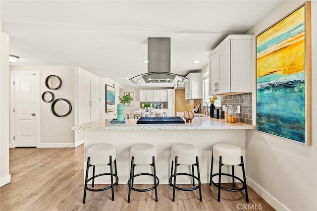 kitchen featuring island range hood, a peninsula, a breakfast bar, white cabinetry, and decorative backsplash