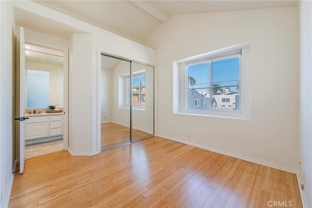 unfurnished bedroom featuring a closet, baseboards, lofted ceiling with beams, light wood-type flooring, and a sink