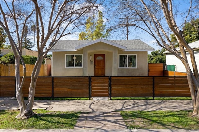 bungalow with a shingled roof, a fenced front yard, and stucco siding