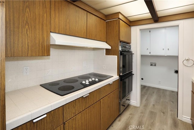 kitchen featuring under cabinet range hood, light wood-style floors, tile counters, brown cabinets, and black appliances