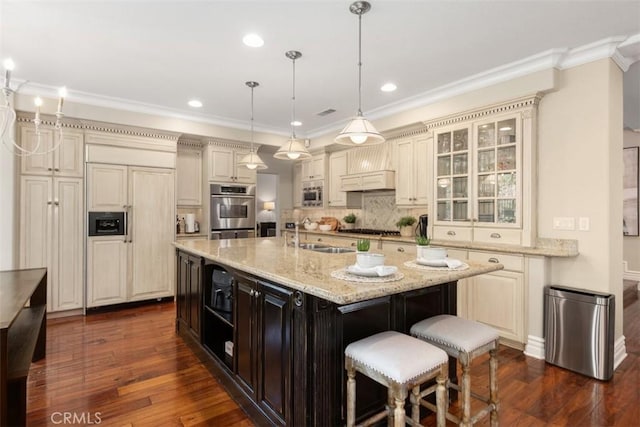 kitchen with built in appliances, light stone counters, a kitchen island with sink, a sink, and glass insert cabinets