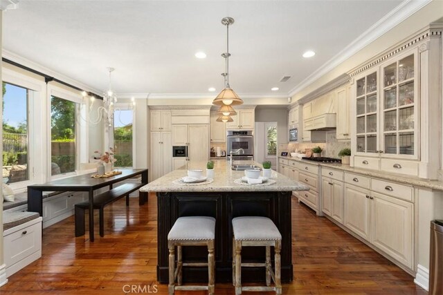 kitchen featuring cream cabinets, glass insert cabinets, stainless steel appliances, hanging light fixtures, and a center island with sink