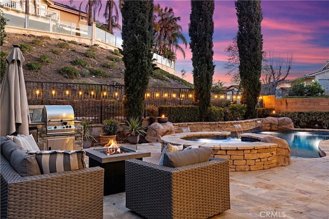 patio terrace at dusk with an outdoor fire pit, a fenced backyard, grilling area, and an outdoor kitchen