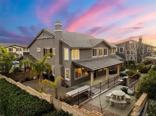 back of house at dusk featuring a patio, a tile roof, a fenced backyard, a chimney, and stucco siding