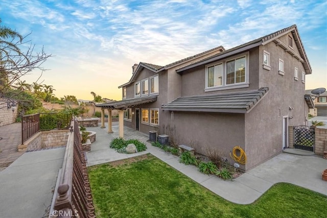 back of house at dusk with fence, a yard, a gate, stucco siding, and a patio area