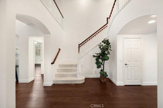 foyer with arched walkways, dark wood-style flooring, a towering ceiling, baseboards, and stairs