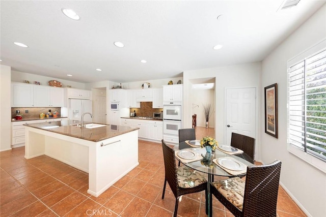 kitchen with light tile patterned floors, a kitchen island with sink, white appliances, a sink, and white cabinets