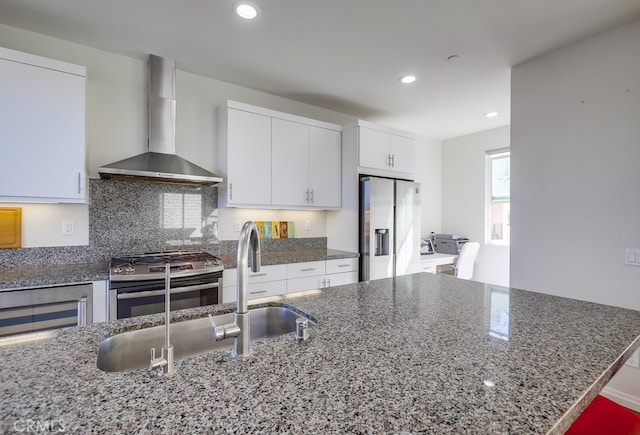 kitchen with stainless steel appliances, wall chimney range hood, dark stone countertops, and white cabinetry