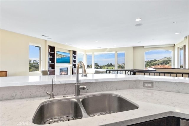 kitchen featuring built in shelves, a sink, open floor plan, light stone countertops, and a glass covered fireplace