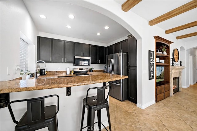 kitchen with appliances with stainless steel finishes, light stone countertops, beamed ceiling, a peninsula, and a kitchen bar