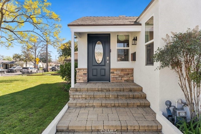 entrance to property with a shingled roof, fence, stone siding, a lawn, and stucco siding