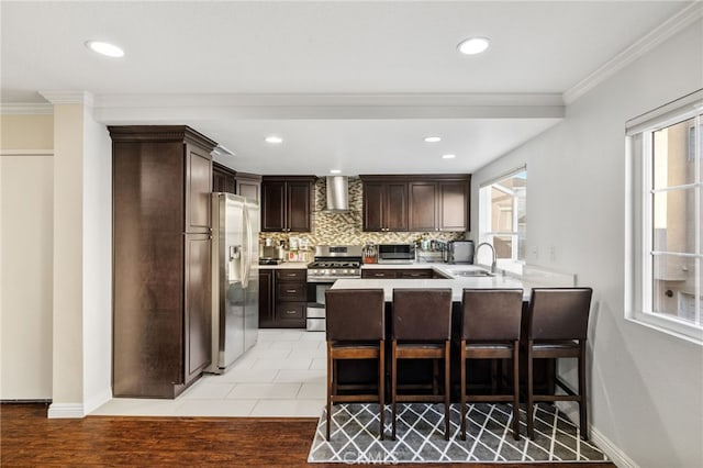 kitchen featuring light countertops, appliances with stainless steel finishes, a healthy amount of sunlight, a peninsula, and wall chimney exhaust hood