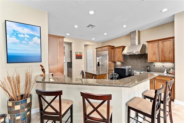 kitchen featuring a breakfast bar area, a peninsula, built in refrigerator, wall chimney range hood, and brown cabinets