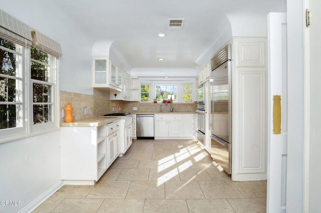 kitchen with visible vents, glass insert cabinets, built in appliances, white cabinetry, and backsplash