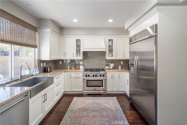 kitchen featuring high quality appliances, glass insert cabinets, light stone countertops, custom exhaust hood, and white cabinetry