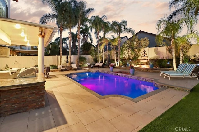 pool at dusk with a patio area, a fenced backyard, and an outdoor kitchen