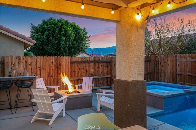 patio terrace at dusk with a fire pit, an in ground hot tub, fence, and a mountain view