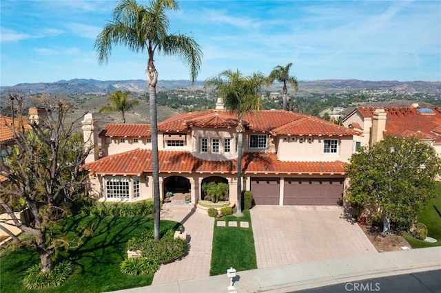 mediterranean / spanish-style home featuring decorative driveway, a tile roof, stucco siding, an attached garage, and a mountain view