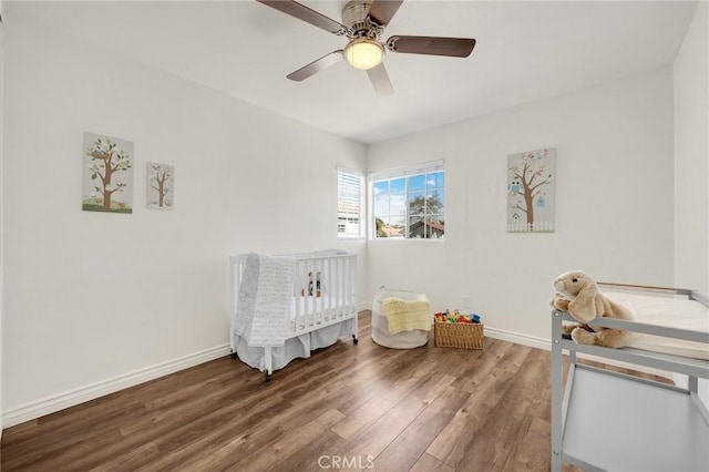 unfurnished bedroom with a nursery area, wood finished floors, a ceiling fan, and baseboards