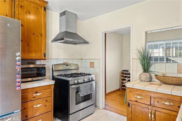 kitchen featuring tile countertops, appliances with stainless steel finishes, brown cabinets, and range hood