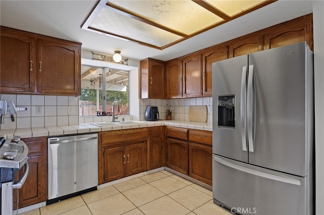 kitchen with tile countertops, light tile patterned floors, stainless steel appliances, a sink, and brown cabinets