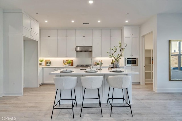 kitchen with a kitchen island with sink, light countertops, under cabinet range hood, white cabinetry, and stainless steel oven