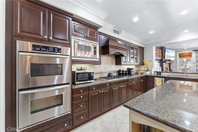 kitchen with light tile patterned floors, dark stone countertops, custom exhaust hood, stainless steel appliances, and a sink