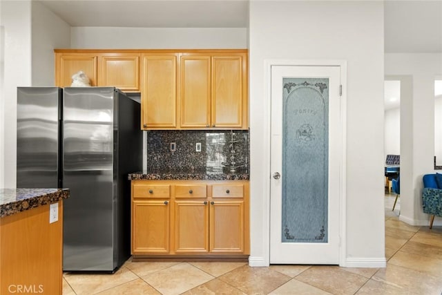 kitchen featuring light tile patterned floors, baseboards, freestanding refrigerator, dark stone counters, and tasteful backsplash