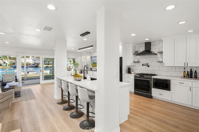 kitchen featuring light wood finished floors, gas range, a breakfast bar, wall chimney range hood, and white cabinetry