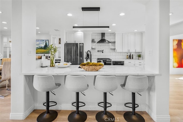 kitchen with wall chimney exhaust hood, a kitchen breakfast bar, stainless steel appliances, light wood-type flooring, and white cabinetry