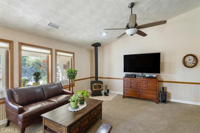 carpeted living room with a wood stove, visible vents, a textured ceiling, and wainscoting