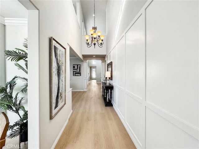 corridor with a decorative wall, a towering ceiling, an inviting chandelier, light wood-style floors, and baseboards