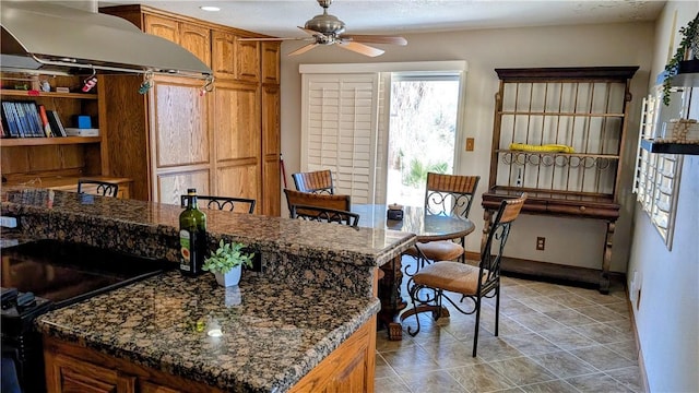 kitchen featuring baseboards, brown cabinetry, dark stone counters, ceiling fan, and a center island