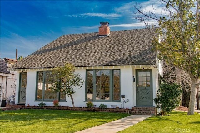 view of front facade with entry steps, a chimney, roof with shingles, a front lawn, and stucco siding