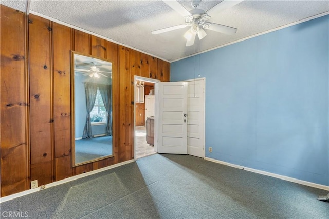 carpeted spare room with a textured ceiling, ceiling fan, wood walls, baseboards, and ornamental molding