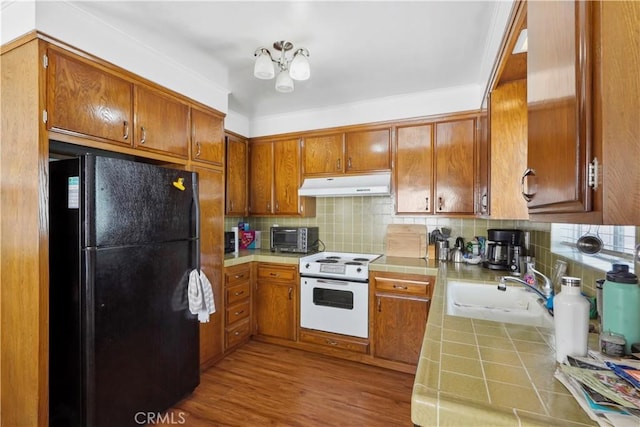 kitchen with white electric stove, under cabinet range hood, tile counters, freestanding refrigerator, and brown cabinetry