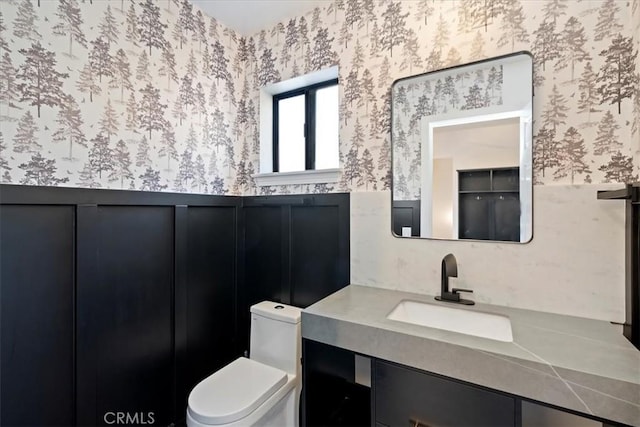 bathroom featuring toilet, wallpapered walls, a wainscoted wall, and vanity