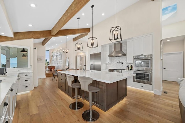 kitchen featuring light countertops, appliances with stainless steel finishes, beam ceiling, wall chimney exhaust hood, and light wood finished floors