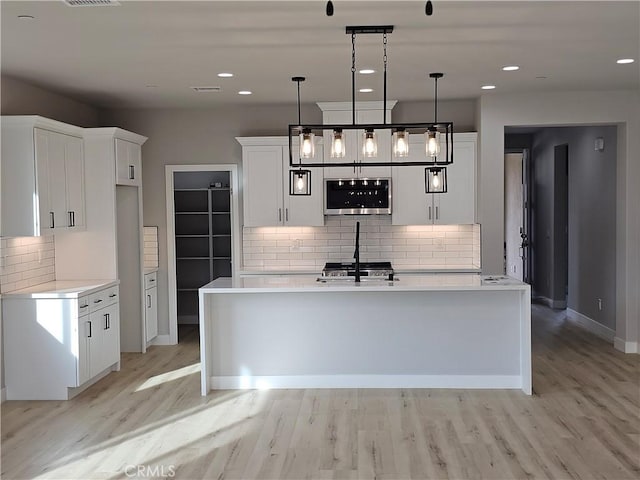 kitchen featuring an island with sink, decorative light fixtures, light countertops, light wood-type flooring, and white cabinetry