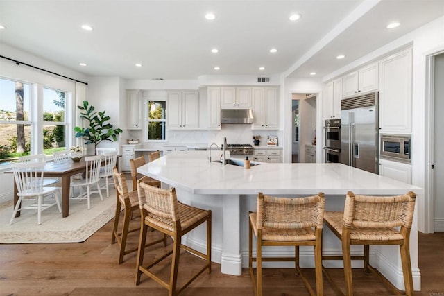 kitchen with tasteful backsplash, wood finished floors, built in appliances, light countertops, and under cabinet range hood