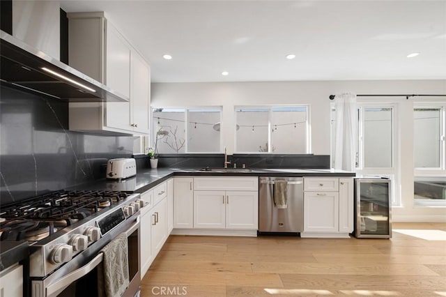 kitchen with wine cooler, stainless steel appliances, light wood-style flooring, a sink, and wall chimney exhaust hood