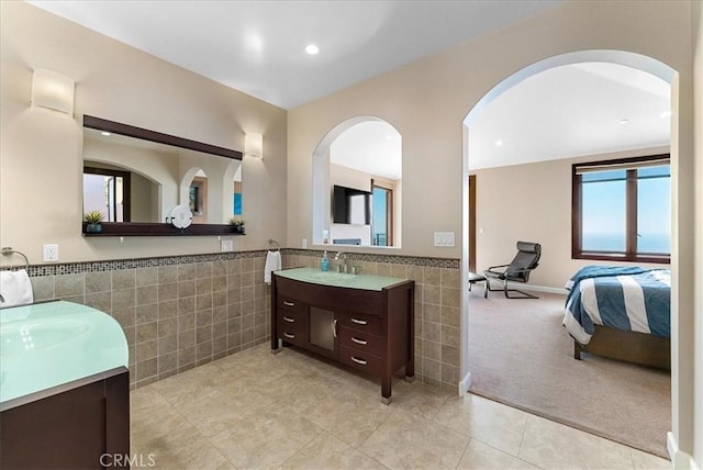 ensuite bathroom featuring tile walls, two vanities, wainscoting, a sink, and ensuite bath