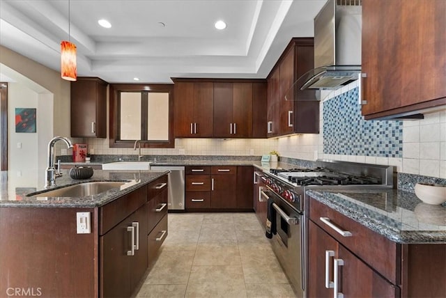 kitchen featuring a raised ceiling, dark stone counters, appliances with stainless steel finishes, wall chimney range hood, and a sink