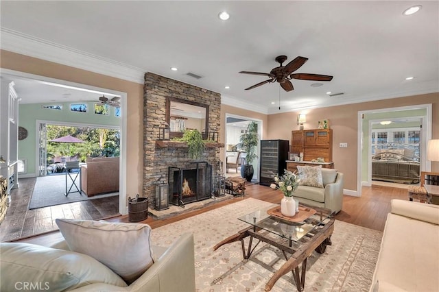 living area with ceiling fan, a stone fireplace, visible vents, light wood-style floors, and ornamental molding