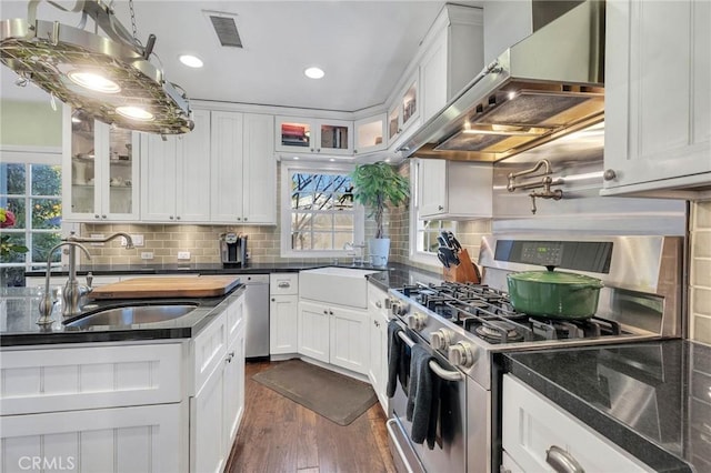 kitchen featuring appliances with stainless steel finishes, glass insert cabinets, white cabinetry, a sink, and wall chimney exhaust hood