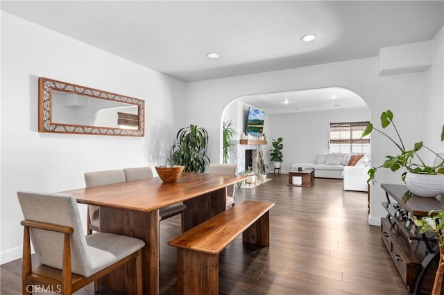 dining room featuring arched walkways, recessed lighting, a fireplace, baseboards, and dark wood finished floors