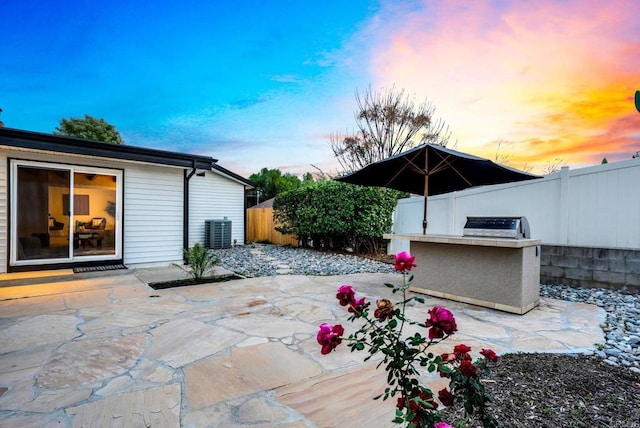 patio terrace at dusk featuring a fenced backyard, an outdoor kitchen, and central air condition unit