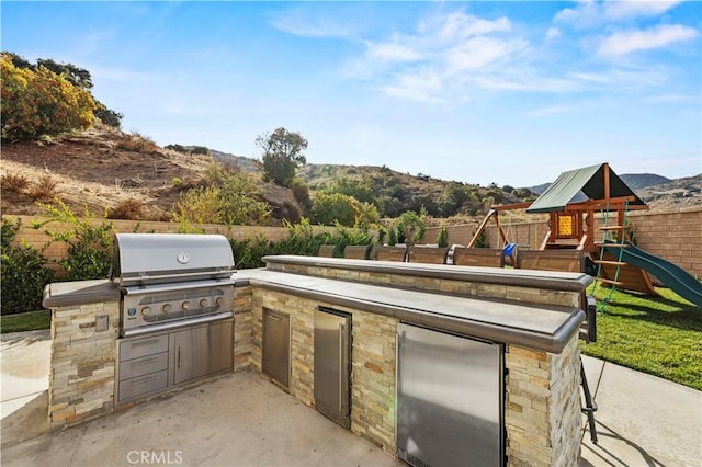 view of patio with exterior kitchen, a playground, fence, and area for grilling