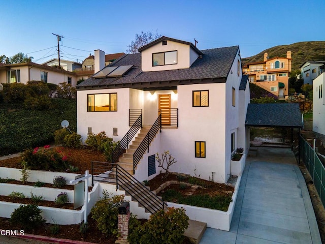 view of front facade featuring roof with shingles, stucco siding, roof mounted solar panels, fence, and stairs